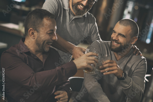 Company of three friends clinking crystal glasses, laughing and looking at each other. Men spending time together in bar, communicating and talking. Friends drinking delicious alcohol.