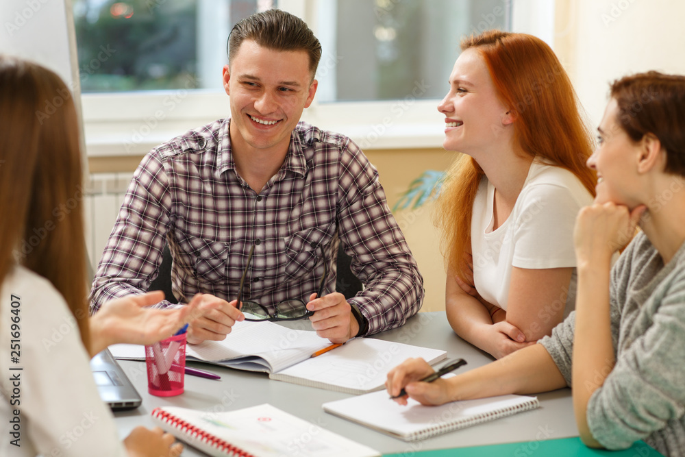 Obraz premium Group of young students sitting together at table and discussing interesting topic in modern school. Handsome boy and pretty girls laughing and enjoying lesson. Concept of education and study.