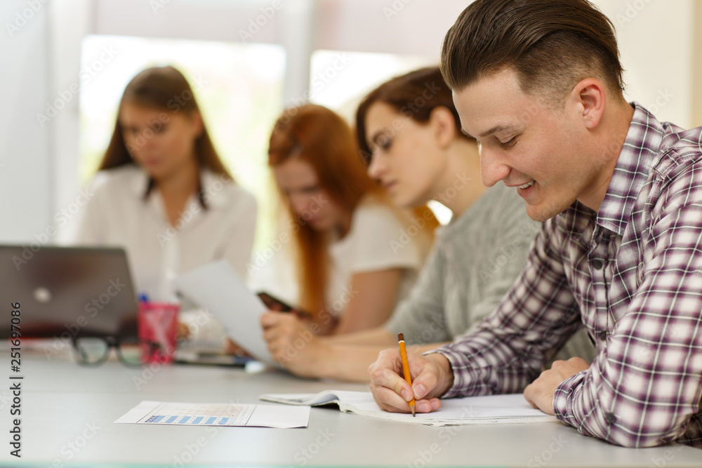 Side view of happy student writing in notes and doing test during lesson in modern school. Young man in checkered shirt smiling because of knowing answers. Concept of interesting studying. Side view of happy student writing in notes and doing test during lesson in modern school. Young man in checkered shirt smiling because of knowing answers. Concept of interesting studying.