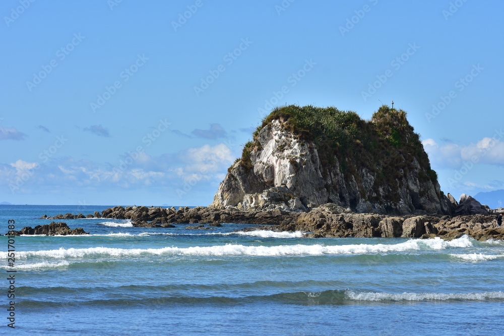 Rocky islet surrounded by flat reef bathing in oceanic surf near mouth of Mangawhai Harbour.