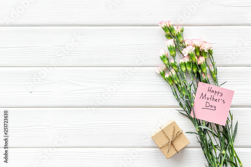 Mother's Day congratulations. Carnations and gift box on white wooden background top view copy space