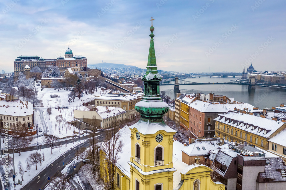 Fototapeta premium Budapest, Hungary - Catholic church with snowy Buda district, Buda Castle Royal Palace, Varkert Bazaar, Szechenyi Chain Bridge and Parliament at background on a snowy winter morning