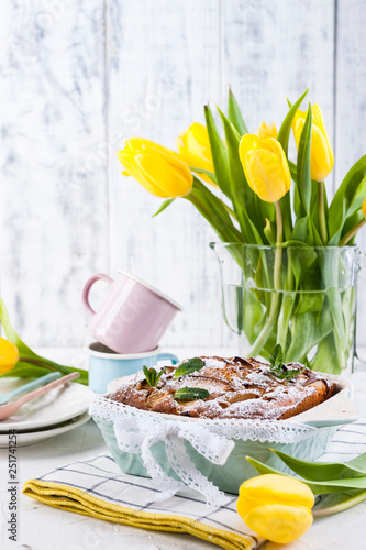Apple pie in dishes on a white background and a bouquet of spring yellow tulips. Homemade pastries with fruit for a delicious breakfast Easter breakfast. Free space for text. Copy space