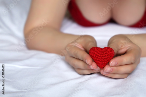 Sexy woman with red knitted heart in hands, declaration of love. Seductive girl in red bra lying on the bed, concept of chastity, romantic sex date, night of love, health care or blood donation