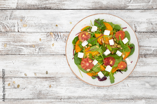 Salad (arugula, chard, mustard salad, spinach, persimmon, red orange, feta cheese, roasted pumpkin seeds, balsamic sauce cream) on white wooden background, top view