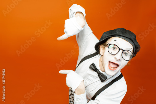 Emotional pantomime with white facial makeup showing empty space on the orange background, advertising something