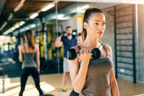 Close up of gorgeous Caucasian brunette doing fitness exercises with dumbbell. In background her reflection in mirror.