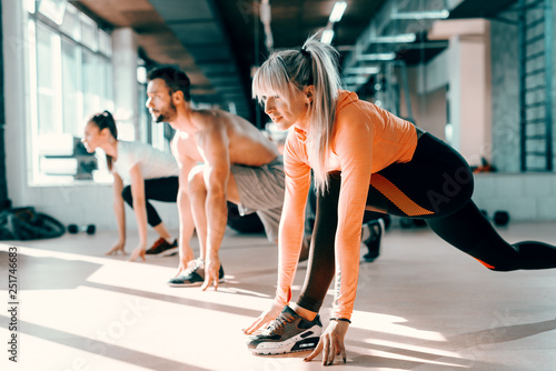 Fototapeta Naklejka Na Ścianę i Meble -  Small group of people with healthy habits doing stretching exercises on a gym floor. Selective focus on blonde woman. In background mirror.