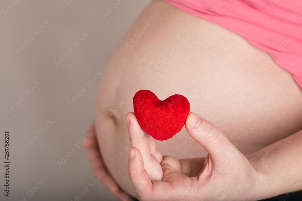 Young pregnant woman keeps small red plush heart close to her belly.
