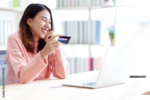 Young happy attractive asian female student, business owner, entrepreneur or freelancer smiling and sitting at home office behind computer laptop holding credit card in hand enjoy shopping online.