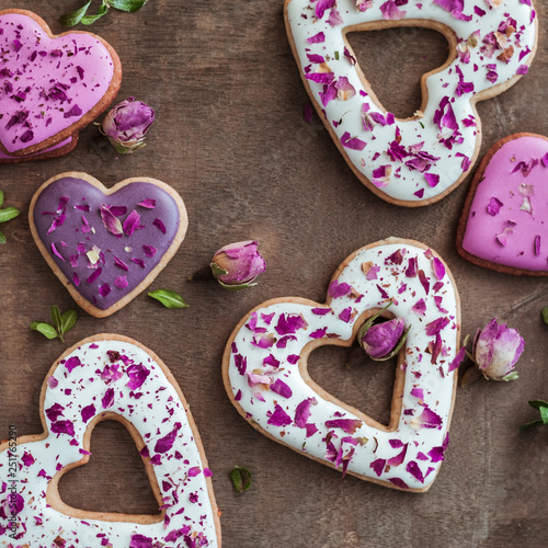 Festive cookies in the shape of hearts and sprinkled with dark pink petals and rosebuds. On a background of brown-gray plywood. Soft focus on petals. Treats and decorations for Valentine's Day. photo