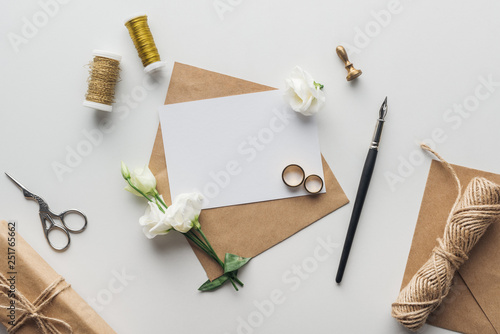 top view of envelope with empty card, ink pen, flowers, stamp, wrapped gift, scissors, spools of thread and golden wedding rings on grey background photo