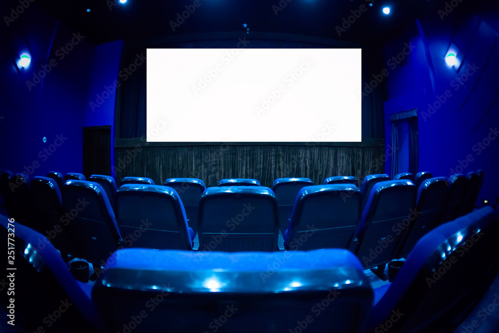 Empty cinema auditorium with empty white screen. Empty rows of theater or movie seats. Blue toned.