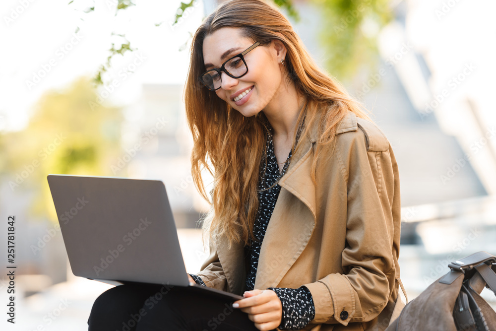 Photo of lovely woman working on laptop while sitting on bench in city street Photo of lovely woman working on laptop while sitting on bench in city street