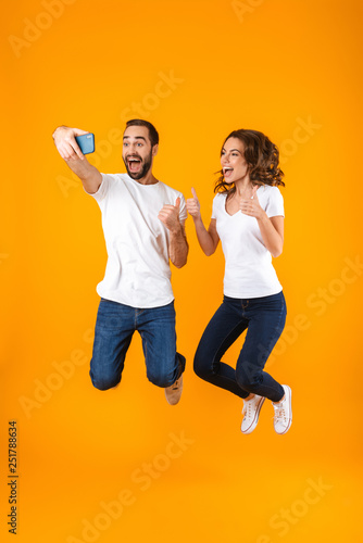 Full length photo of content couple laughing and taking selfie on smartphone, isolated over yellow background