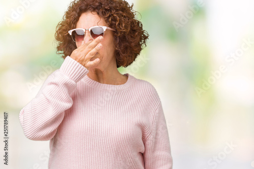 Beautiful middle ager senior woman wearing pink sweater and sunglasses over isolated background smelling something stinky and disgusting, intolerable smell, holding breath with fingers on nose