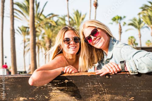 Cheerful couple of young beautiful caucasian girls smiling and looking at the camera - braket for perfect teeth and friendship for happy people outdoor in summer time - vacation and tropical palms photo