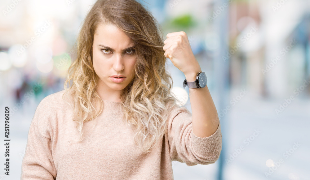 Beautiful young blonde woman wearing sweatershirt over isolated background angry and mad raising fist frustrated and furious while shouting with anger. Rage and aggressive concept.