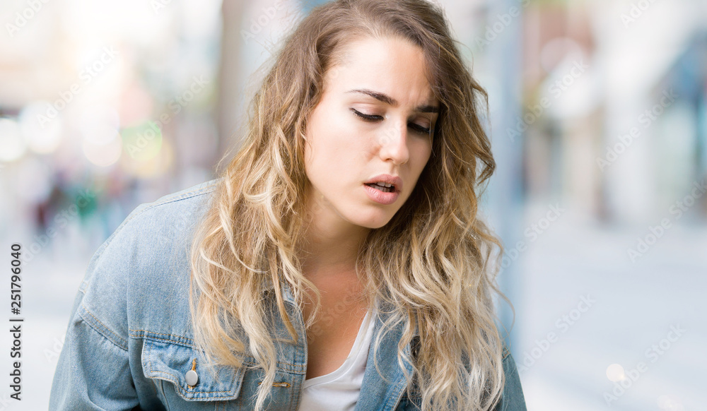 Beautiful young blonde woman wearing denim jacket over isolated background with hand on stomach because nausea, painful disease feeling unwell. Ache concept.