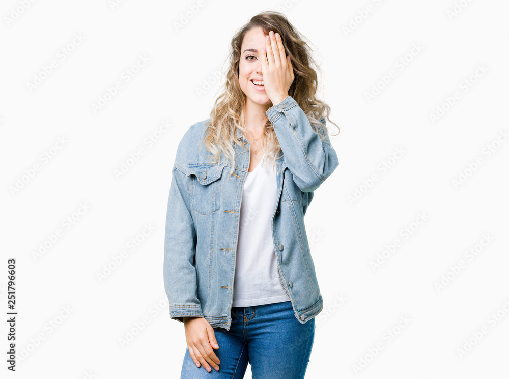 Beautiful young blonde woman wearing denim jacket over isolated background covering one eye with hand with confident smile on face and surprise emotion. Beautiful young blonde woman wearing denim jacket over isolated background covering one eye with hand with confident smile on face and surprise emotion.
