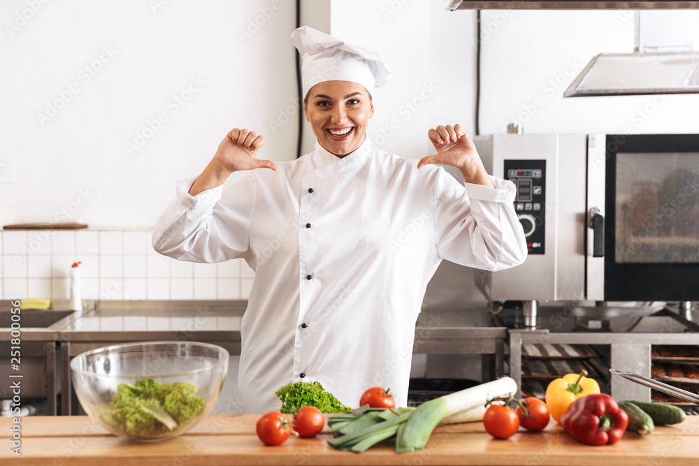 Photo of smiling woman chef wearing white uniform cooking meal with fresh vegetables, in kitchen at the restaurant Photo of smiling woman chef wearing white uniform cooking meal with fresh vegetables, in kitchen at the restaurant