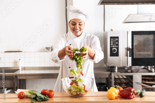 Photo of pleased woman chef wearing white uniform making salad with fresh vegetables, in kitchen at the restaurant