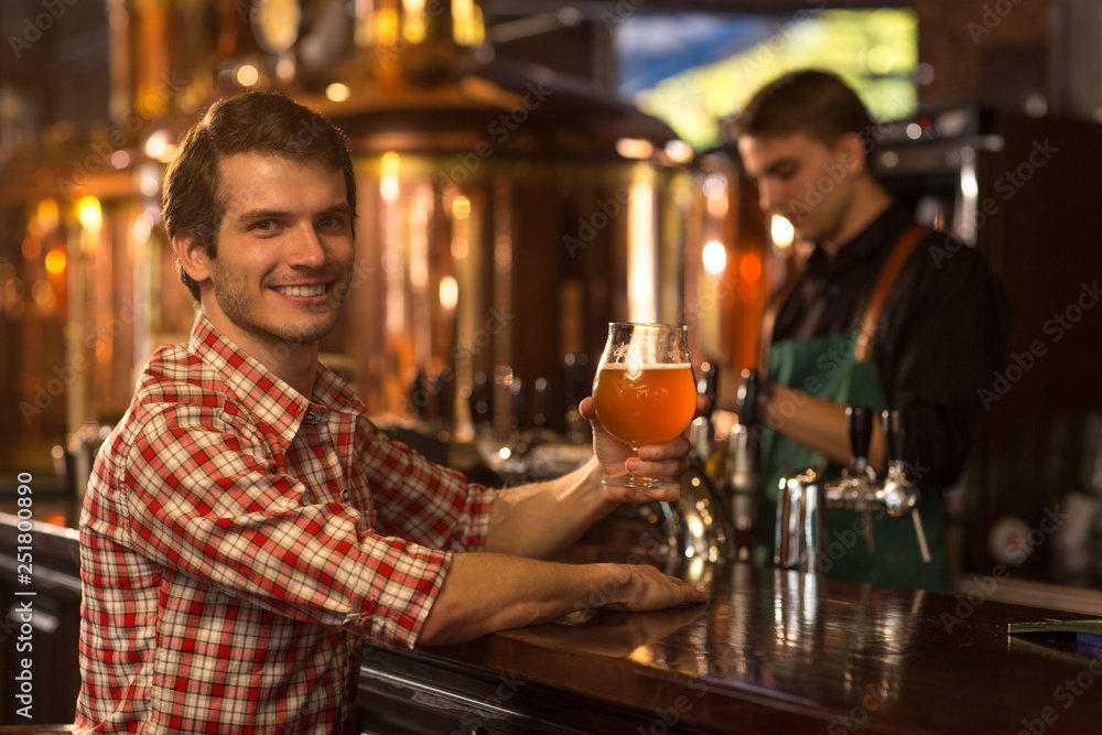 Handsome man in checked shirt sitting at bar counter and holding glass of beer. Client of brewery looking at camera, smiling and posing. Barmen working and bronzed kettles behind.