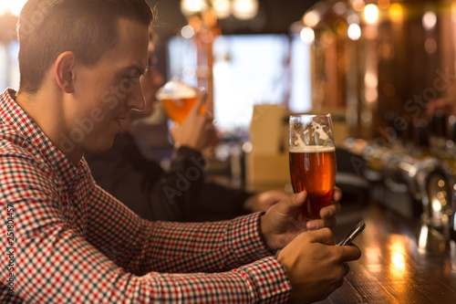 Side view of client of bar sitting at bar counter  holding beer glass in hand. Man smiling and looking at mobile phone. Man spending time in beer house or brewery  tasting delicious beer and resting.