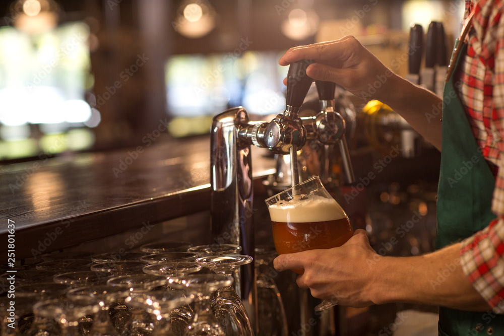 Hands of unrecognizable bartender holding glass and filling it with delicious fresh beer. Barman working at bar counter with alcohol beverages in beer house or brewery.
