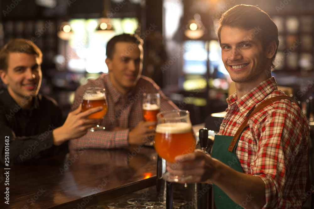 Positive bartender posing, smiling and showing beer. Handsome barman wearing in checked shirt and apron. Clients of beer house or brewery sitting with beer glasses in hands behind.