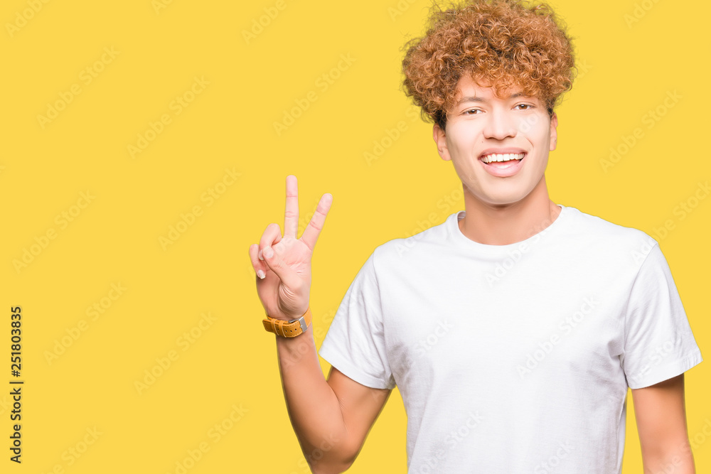 Young handsome man with afro hair wearing casual white t-shirt smiling with happy face winking at the camera doing victory sign. Number two. Young handsome man with afro hair wearing casual white t-shirt smiling with happy face winking at the camera doing victory sign. Number two.