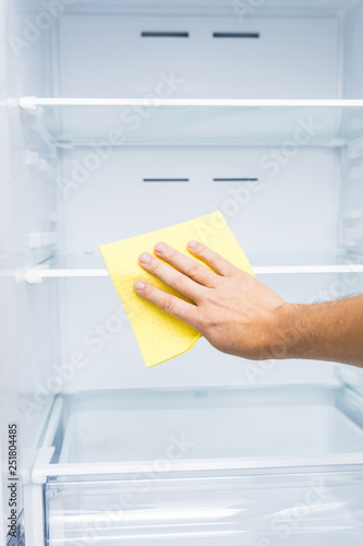 Man hand in yellow rubber protective glove cleaning white open empty refrigerator with green rag