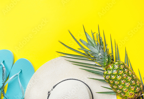 Ripe pineapple on green palm leaf white straw hat blue slippers on bright yellow solid background. Summer vacation fun tropical fruits beach party concept. Trendy flat lay