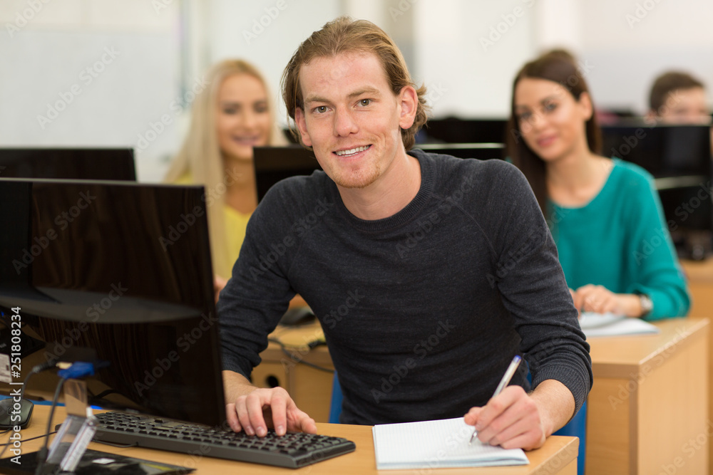 Handsome young man sitting at table, using personal computer and writing lecture in notebook. Smart student wearing in casual clothes, smiling and looking at camera at university. Education concept. Handsome young man sitting at table, using personal computer and writing lecture in notebook. Smart student wearing in casual clothes, smiling and looking at camera at university. Education concept.