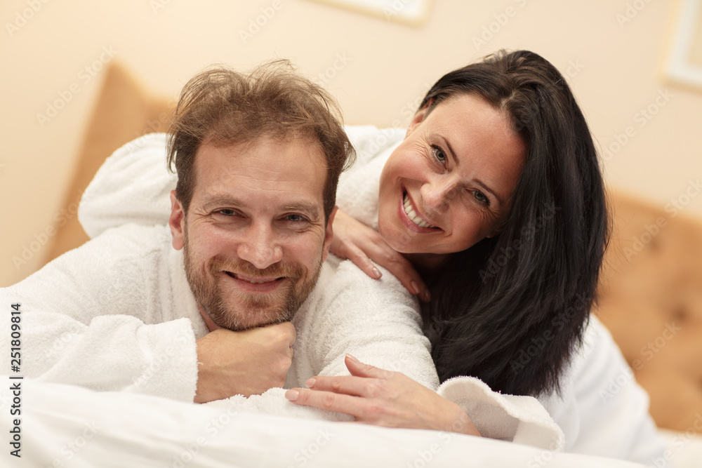 Portrait of laughing couple in white robes lying on bed, looking at camera and posing. Cheerful man and woman resting together and enjoying time spending in morning. Concept of happiness. Portrait of laughing couple in white robes lying on bed, looking at camera and posing. Cheerful man and woman resting together and enjoying time spending in morning. Concept of happiness.