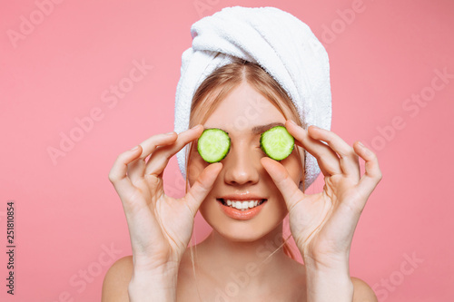 Attractive cheerful woman with a towel wrapped around her head, holding cucumber slices near her eyes, on a pink background