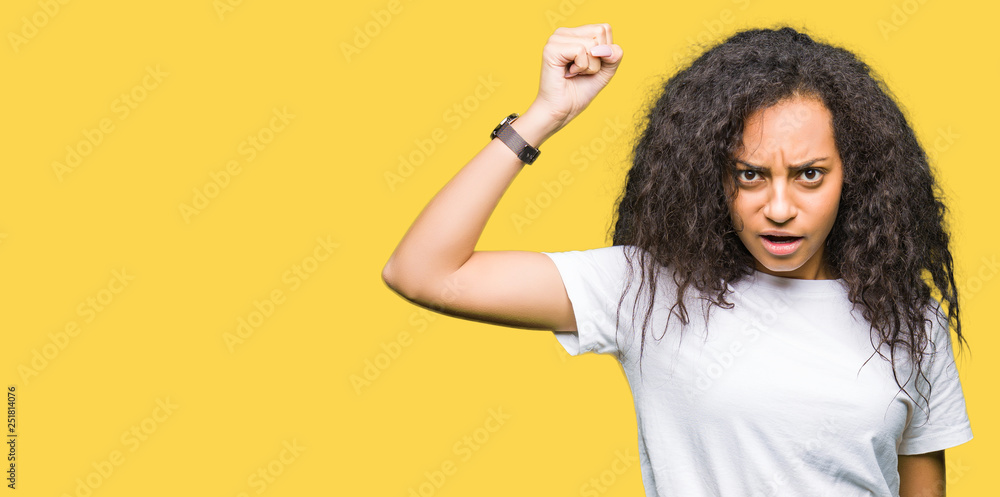 Young beautiful girl with curly hair wearing casual white t-shirt angry and mad raising fist frustrated and furious while shouting with anger. Rage and aggressive concept. Young beautiful girl with curly hair wearing casual white t-shirt angry and mad raising fist frustrated and furious while shouting with anger. Rage and aggressive concept.
