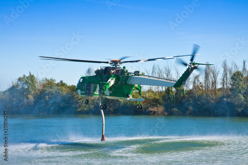 Italian CFS 103 fire fighting helicopter flying over a lake to collects water through a suction tube to extinguish a fire photo