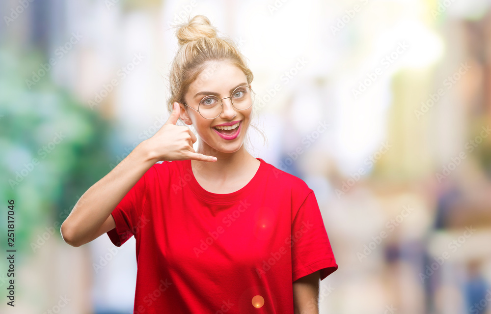 Young beautiful blonde woman wearing red t-shirt and glasses over isolated background smiling doing phone gesture with hand and fingers like talking on the telephone. Communicating concepts.
