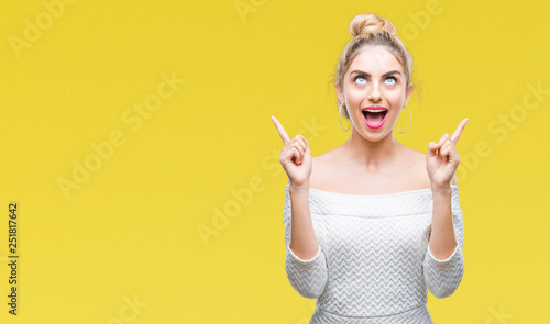Young beautiful blonde and blue eyes woman over isolated background amazed and surprised looking up and pointing with fingers and raised arms.
