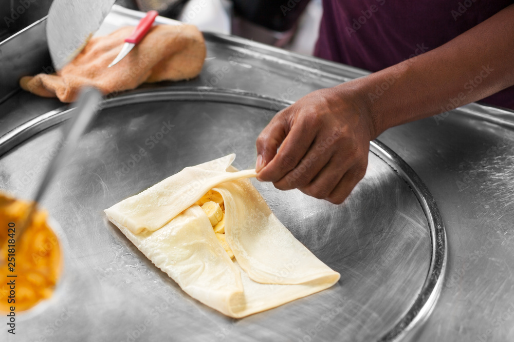 Cooking traditional Thai fried roti banana pancakes close up, asian street food preparation in Thailand, man chef hands frying pancakes on griddle in Bangkok, sweet fruit banana roti making on hot pan
