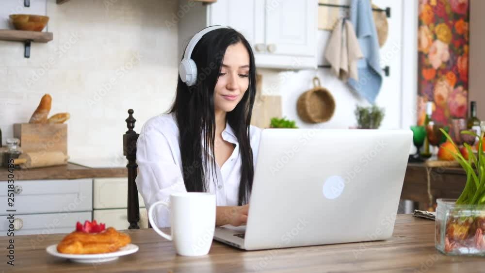 Smiling Young Woman Using Laptop In The Kitchen At Home Wearing Headphones Smiling Young Woman Using Laptop In The Kitchen At Home Wearing Headphones