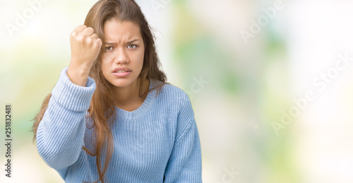 Young beautiful brunette woman wearing blue winter sweater over isolated background angry and mad raising fist frustrated and furious while shouting with anger. Rage and aggressive concept. © Krakenimages.com