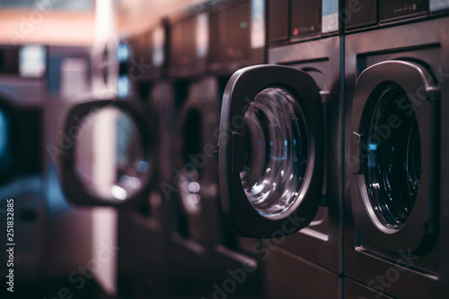 Slika na platnu A dark room of a publiс laundry with a row of empty washing machines inside, sel