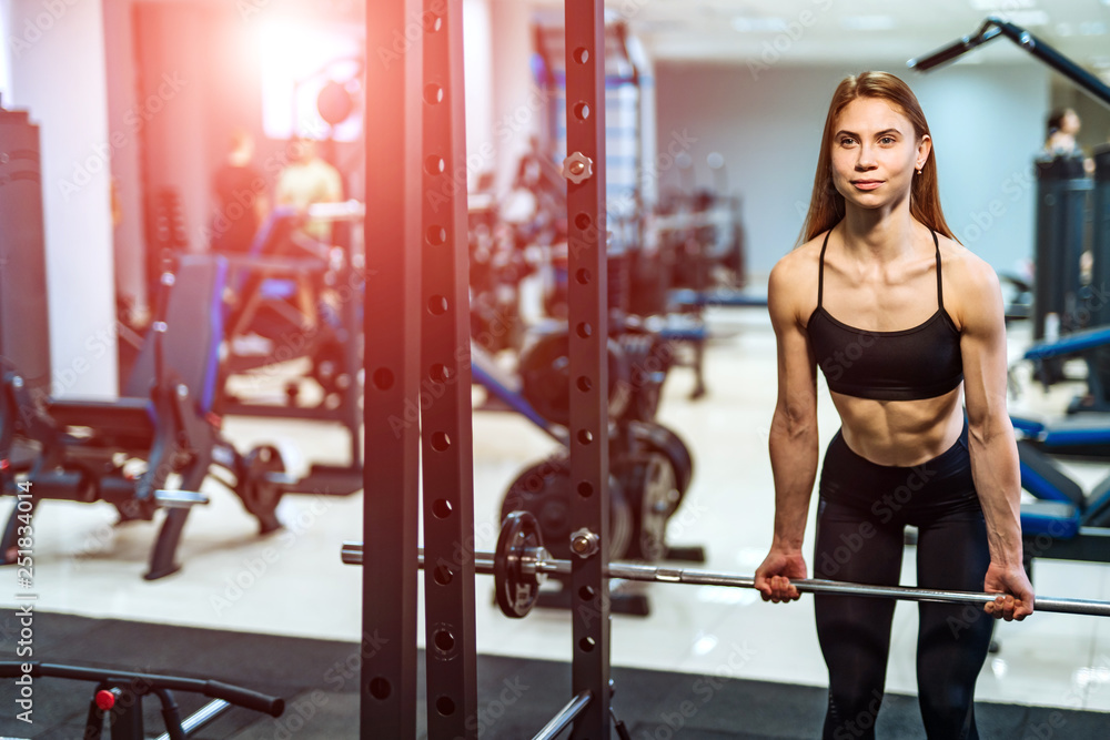 Fitness girl exercising with barbell in gym. Young beautiful fitness girl posing in gym with sports equipment Fitness girl exercising with barbell in gym. Young beautiful fitness girl posing in gym with sports equipment