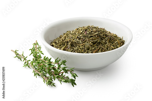 Dried rubbed thyme in a white ceramic bowl next to fresh thyme sprigs isolated on white.