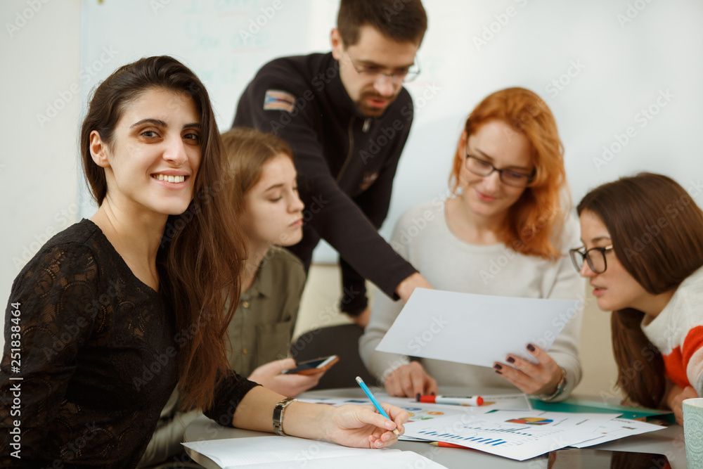 Group of people discussing some topics during lessons. Teacher explaining to students graphics and schematics on examples. Girl smiling at camera while other pupils listening to professor. Group of people discussing some topics during lessons. Teacher explaining to students graphics and schematics on examples. Girl smiling at camera while other pupils listening to professor.