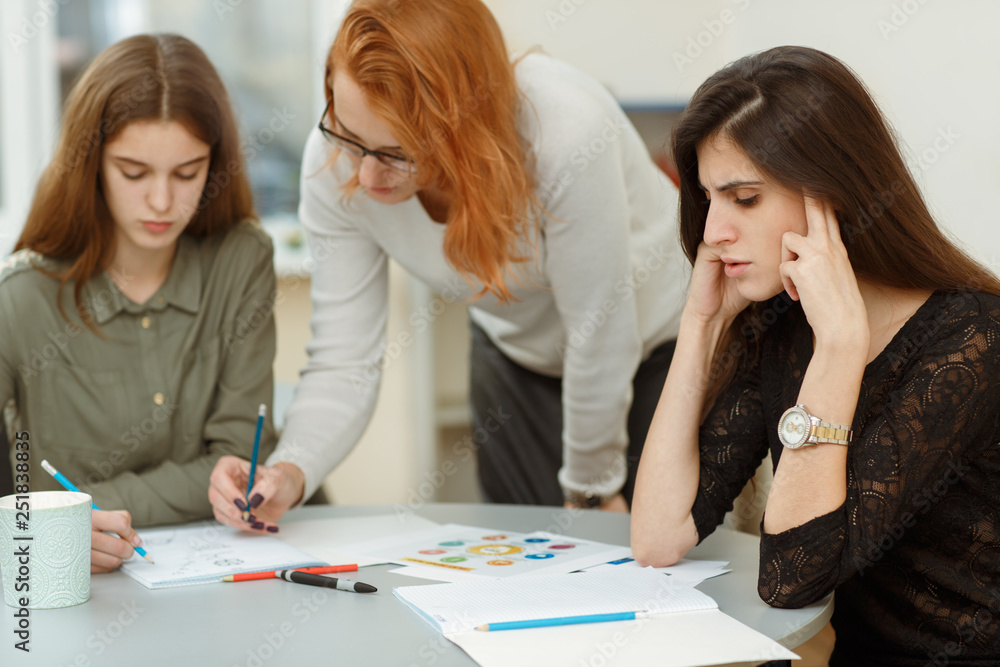 Pupils hardly thinking under difficult tasks during classes. Young teacher helping and explaining graphics and schematics to her students. Red haired tutor checking homeworks of beautiful girls. Pupils hardly thinking under difficult tasks during classes. Young teacher helping and explaining graphics and schematics to her students. Red haired tutor checking homeworks of beautiful girls.