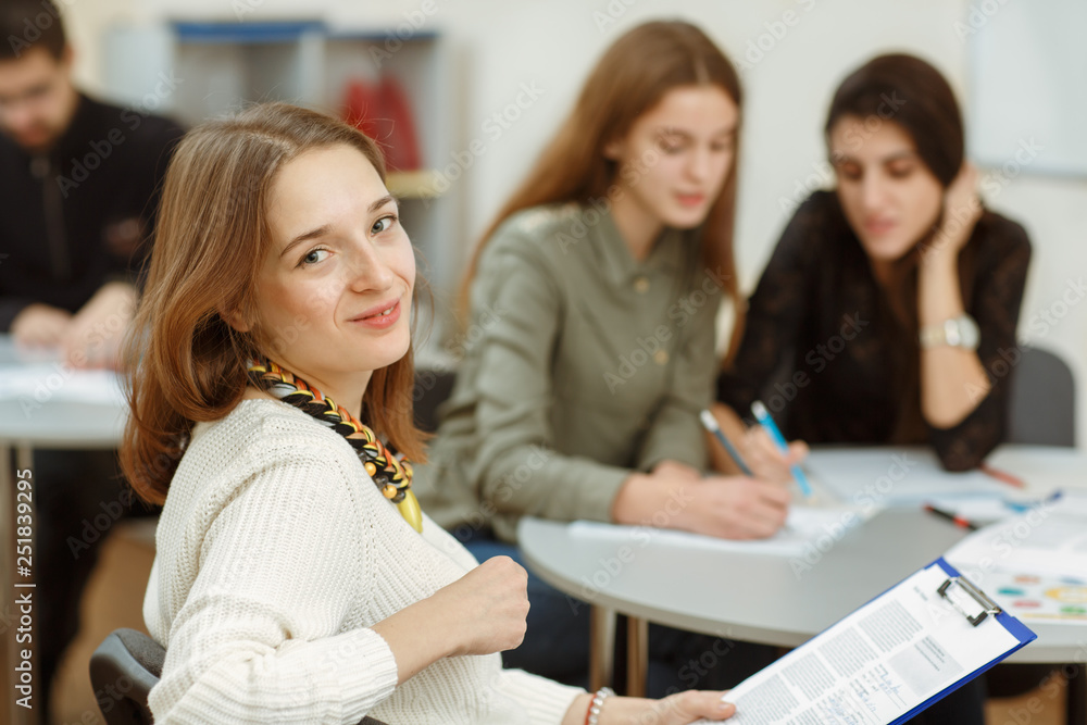 Crop of beautiful young girl sitting in class and looking at camera. Red haired happy student writing down new topic in copybook. On background pupils doing exercises by themselves during lecture. Crop of beautiful young girl sitting in class and looking at camera. Red haired happy student writing down new topic in copybook. On background pupils doing exercises by themselves during lecture.