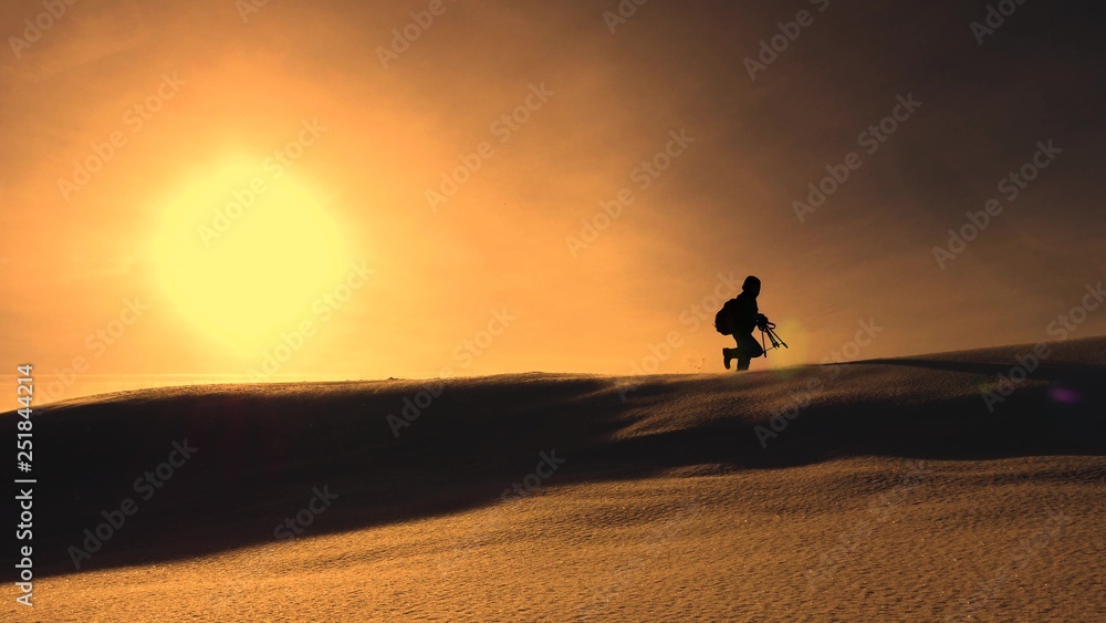 photographer traveler in winter goes on snowy ridge in rays of yellow sunset. mountaineer with camera and tripod is walking in snow along top of holom.